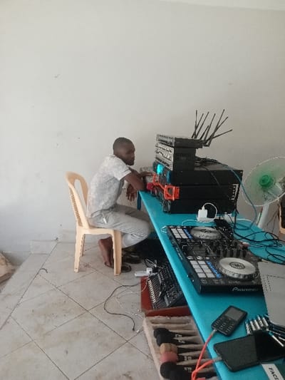 The image displays an indoor scene located in Kinondo ward, Kenya. One dark-skinned adult male, with short hair and a beard, is seated on a light-colored plastic chair. He wears a light-patterned short-sleeved shirt and matching long pants, with bare feet resting on a light-tiled floor. His forearms are positioned on a bright blue table surface, with hands near the equipment.
The table features a stack of black rack-mounted audio equipment, including units with blue LED indicators and a top unit equipped with multiple antennae. An orange plastic bracket supports a section of this stack. To the right, a light green desk fan is present. A black Pioneer brand DJ controller, featuring two jog wheels, buttons, and faders, occupies the table's right side. Adjacent items include a smaller black audio mixer, a black feature phone, and a black smartphone connected by an orange cable. Multiple audio jacks/adapters are situated on the far right of the table. Black and white cables are arranged on both the table and the tiled floor, connecting various devices, with a white power strip visible on the floor. A wooden tray containing several microphones, distinguished by black, pink, and beige foam windscreens, rests on the floor partially beneath the table in the foreground.