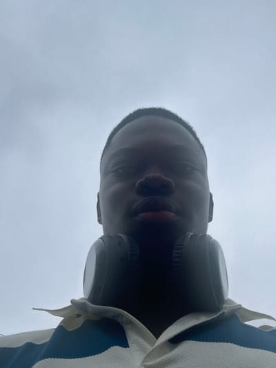 An individual looks upwards towards the camera. Wearing headphones, the person’s face is framed by the overcast sky. The camera angle is low, emphasizing the face against the backdrop. A collared shirt with blue and white stripes is visible on the right side of the frame. The person's expression is neutral.