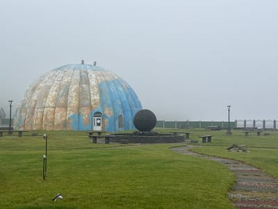 A large, dome-shaped structure resembling a weathered globe sits on a grassy expanse under a thick, foggy sky. The dome, painted with continents in shades of blue and brown, has a single door and two arched windows. In front of the dome is a circular stone fountain with a large, dark, spherical sculpture at its center, surrounded by stone benches. A winding path made of colored stones leads through the manicured grass, where several other stone benches and a solitary lamppost are visible. A small bird in flight is captured near the bottom left of the frame. The overall scene suggests a quiet, possibly abandoned, outdoor space in a location characterized by overcast weather and low visibility due to fog. The time of day is indeterminate due to the uniform, diffused light. The setting appears to be a park or public area, with decorative elements and seating areas. There is no visible text in the image.