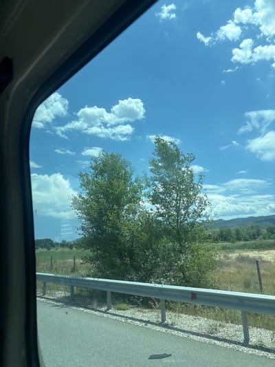 View from inside a vehicle through an open sunroof, looking towards a bright blue sky with scattered white clouds. To the right, roadside vegetation and a metal guardrail are visible. Further in the background, a landscape including fields and distant mountains is seen. The location is Strumyani, Bulgaria.