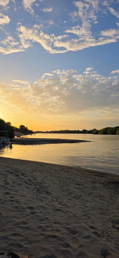 The image captures a serene sunset scene by the Pripyat River in Mazyr, Belarus. The sky is a gradient of soft oranges and yellows, transitioning into a pale blue, with scattered, puffy white clouds. The calm river reflects the warm hues of the sky, creating a shimmering, golden surface.
In the foreground, a sandy beach stretches across the lower portion of the frame. The sand shows subtle textures and ripples. To the left, near the water's edge, a small white boat is docked, with a dark-colored structure, possibly a canopy, above it. Two figures are visible near the boat, one appearing to be standing.
Across the river, a dense line of green trees forms a natural treeline along the opposite bank. Further out on the water, a few scattered dots suggest people engaged in activities on the river or in the water. The overall atmosphere is peaceful and tranquil, characteristic of a late summer evening by the water.