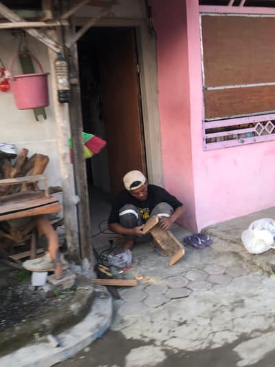 A man, wearing a black t-shirt, grey pants, and a beige baseball cap, is crouched on a concrete pathway in Keboguyang, Indonesia. He is working on a piece of wood, holding a rasp or file in his right hand and the wood in his left. A power tool is visible on the ground near his feet, along with a broom with red, green, and yellow bristles. To his right, a white plastic bag and a pair of purple flip-flops are on the ground. A pink building with a brown window is to his right, and a wooden structure with various objects, including a pink bucket, is to his left. The ground is a mix of concrete and hexagonal paving stones, with some debris and a water channel in the foreground.