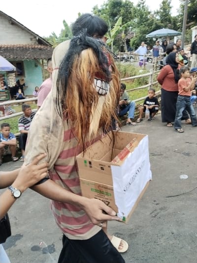 The image depicts a daytime scene on a paved road in the city of Sumberejo, Indonesia, during what appears to be a local event or procession.

In the foreground and center, a person is walking forward, viewed from the right side. This individual is wearing an elaborate, unkempt mask featuring a white face with a prominent black-toothed grimace, and long, wet-looking strands of black, brown, and blonde hair. They are dressed in a dirty, light-colored long-sleeve outer shirt over a red and beige striped t-shirt, black pants, and light beige slide sandals. The person is carrying a multi-compartment cardboard box with a white sheet of paper taped to its front, bearing partially visible vertical text "SEIKHONY...". An arm, belonging to an unseen individual, is visible on the left, grasping the masked person's upper left arm/shoulder area. This arm wears a silver watch on the wrist.

The background shows a mix of residential structures and natural elements. To the immediate left, a house with a tiled roof and light blue walls is visible, alongside a small vendor stall with a purple umbrella. Several onlookers, including adults and children, are seated or standing near this house. To the right, a crowd of approximately ten to fifteen adults and children observes the scene. Some individuals are standing on the paved road, while others are positioned behind a rustic wooden fence that separates the road from a lower grassy area. Trees and dense green foliage are visible further in the background, along with additional distant houses. The road surface is light gray asphalt, exhibiting some dark stains. The sky is bright but overcast, indicating diffused daylight.