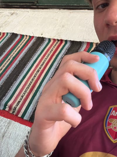 A person holds a blue microphone close to their mouth. The person is wearing a maroon shirt with a yellow and red patch on the upper right chest area and a silver bracelet on their right wrist. A striped rug with shades of gray, green, red, white, and black is partially visible. A white textured wall and a green door are partially visible on the left. The location is not identifiable as Kanchevo, Bulgaria from the visual information provided.