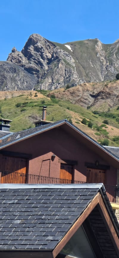 The image shows a close-up view of a building with a dark slate roof, situated against a backdrop of rugged, green mountains and a clear blue sky. The building's exterior is a warm, reddish-brown color, with dark wooden shutters or doors visible. A metal chimney pipe extends from the building. The mountains are rocky and steep, with patches of green vegetation and some remaining snow on the higher slopes. The sunlight suggests it is daytime, and the clear sky indicates fair weather. The scene evokes a sense of remoteness and natural beauty, typical of a mountain landscape. The image appears to be taken from a slightly elevated perspective, looking down at the building and the surrounding terrain. There is no visible text in the image.