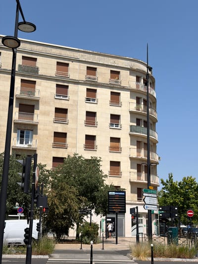 This is a street-level, medium shot of a building and intersection in Courbevoie, France, on a clear, sunny day. The building is a light-colored, multi-story structure with rounded corners and a facade of stone blocks. It has numerous windows, many with closed brown shutters, and several balconies with white railings. A tall black lamppost with modern lighting fixtures stands prominently in the foreground to the left.

In the intersection, a crosswalk is visible on the pavement. Traffic lights are present, with one showing a red pedestrian signal (a standing figure). A white van is parked near the curb on the left. Several signs are mounted on poles. One sign displays "Informations" and lists times and days, possibly related to business hours. Another set of green directional signs indicate "LA DEFENŠE", "Gare S.N.C.F. de Courbevoie", and "CHARRAS". A person is standing next to a bicycle near the entrance to a building with a sign that reads "CORBEIL". Trees with green foliage are scattered throughout the scene, some obscuring parts of the building and signs. The sky is a clear, bright blue.