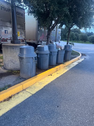 The image depicts a row of five grey plastic trash cans, each with a lid, lined up next to a yellow-painted curb. The trash cans are situated on a paved surface, likely a parking lot or roadside, with a concrete structure to the left. In the background, several large semi-trailer trucks are parked. One truck has "Hurricane Express" and "Recruiting" printed on its side. Trees with green foliage are visible above and behind the trucks. The scene appears to be outdoors, with a natural, park-like setting visible further in the background. The lighting suggests it is either early morning or late afternoon. The location is identified as Lake City, United States.
