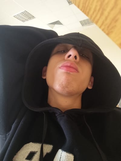 A close-up, upward-angled image depicts an individual, presumed male, wearing a black hooded sweatshirt. The hood is pulled over the head, partially obscuring the forehead and the upper portion of the eyes, casting shadows. The visible facial features include a prominent nose, full lips with a slight pout, and light stubble on the chin and jawline. The individual is looking directly towards the camera. A portion of a light-colored, textured applique, possibly lettering, is visible on the chest of the sweatshirt on the viewer's left. The individual's left arm, bent at the elbow, is visible on the left side of the frame. The background consists of an interior ceiling featuring white panels, several square recessed lighting fixtures, and at least one rectangular ventilation grille. A light-colored wall or surface is visible on the upper right. The specific location within Jibb as-Safa, Syria, cannot be determined from the provided visual information.