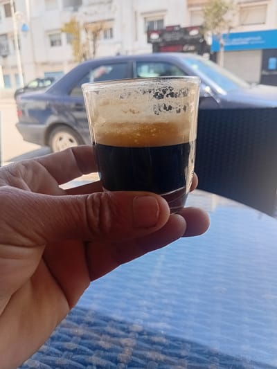 A hand holds a small glass of coffee, likely espresso, with a layer of crema on top. The scene is outdoors on a blue, patterned table, potentially at a café. A car is visible in the blurred background, parked on a street with buildings. The location is Martil ⵎⴰⵔْتⵉⵍ مرتيل, Morocco, suggested by the architectural style of the buildings and the overall ambiance.