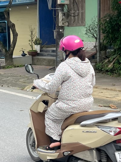 A person wearing a pink helmet and a floral-patterned outfit is riding a beige scooter. The scooter is on a street in Hà Giang 2 Ward, Vietnam. The background shows a building with a yellow shuttered storefront, stairs, and lush greenery. The street appears to be paved, with a concrete curb and some fallen leaves. The overall scene depicts an everyday street view in a Vietnamese urban setting.