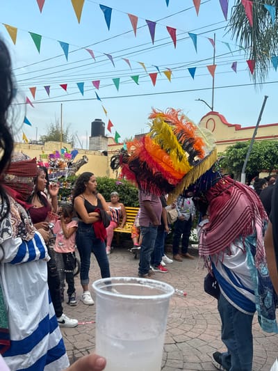 The image captures a festive scene in Oacalco, Mexico. In the foreground, a large plastic cup is held up. Behind it, a gathering of people is visible on a paved area. The attire of some individuals, including vibrant feather headdresses, suggests a cultural event or parade. A woman in a black top stands with her arm around a child, looking towards the spectacle. Another woman is seated, possibly taking a break. Decorative triangular flags are strung overhead, adding to the ambiance. In the background, there's a building with a light-colored facade and a water tank on its roof, further defining the location.