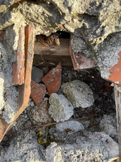 The image depicts a close-up view of a hole in a weathered, possibly abandoned, structure. The hole reveals a jumble of debris, including broken pieces of reddish-orange ceramic tiles, fragments of concrete or plaster, and what appears to be a piece of aged, splintered wood. Cobwebs are visible within the opening, suggesting disuse. The surrounding material is rough and textured, with areas of lighter gray and darker gray, indicating the passage of time and exposure to elements. The lighting suggests it is daytime, with sunlight casting shadows and highlighting the textures. The scene is one of decay and neglect. The specific location in Podostrog, Montenegro, is not evident from the visual information alone, but the context of a hole in a structure with debris suggests an outdoor, possibly rural or semi-rural setting.