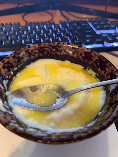 A metallic spoon rests in a brown ceramic bowl filled with melted butter and what appears to be cooked eggs. The bowl has a speckled, textured pattern around its rim. In the background, a keyboard with blue backlighting is visible, suggesting the scene is indoors, likely in a home or office. No specific location in Tula, Russia can be identified.