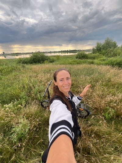 A medium shot shows a woman taking a selfie in a grassy field near the city of Pivkino, Russia. The woman, with her hair pulled back, is smiling at the camera and her right hand is extended, palm open, towards the right. She is wearing a black and white letterman jacket. Behind her, a bicycle is lying on its side in the tall grass. The background features a body of water, likely a river, and a distant treeline with some buildings visible. The sky above is filled with dramatic, dark clouds, with a sliver of sunlight breaking through on the horizon.