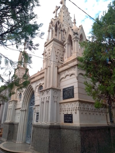 The image shows a mausoleum with Gothic architectural elements, featuring a cross at the top. There are trees partially obscuring the view of the structure. A plaque is visible on the side of the mausoleum with the inscription "Gral. de Brig. E. ANDRES RODRIGUEZ," commemorating the date of February 2 & 3. The location is likely in a cemetery in Asuncion, Paraguay.