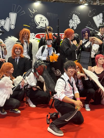 At the Paris Nord Villepinte Exhibition Centre in Villepinte, France, a group of cosplayers, likely attending a convention, are posing for a photograph on a bright red carpet. The backdrop features a black wall with white graphic designs, possibly related to a manga or anime series. The individuals are dressed in costumes, many of whom have orange hair and appear to be portraying characters from "Chainsaw Man," including Denji (with a chainsaw head), Aki Hayakawa, and possibly Power. Some are wearing formal attire like suits and ties, with blood splatters on some of their white shirts. One person is holding a plush toy of Pochita, the chainsaw dog. Another individual is brandishing a prop sword. Several people are wearing white feathered wings. The lighting suggests an indoor convention environment.