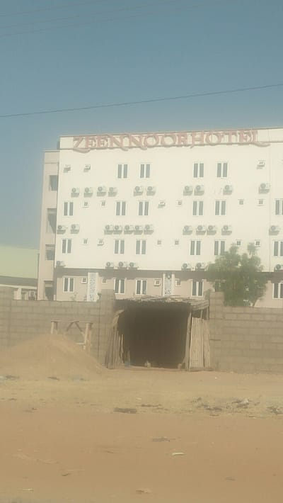 A dusty foreground leads to a walled construction site, revealing a dimly lit, tunnel-like opening. Behind it stands the Zeennoor Hotel, a tall building with rows of windows and air conditioning units. A few trees are visible on the right side of the construction site, and power lines stretch across a clear sky above the hotel.