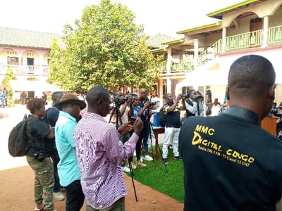 A group of about 15 people stand in the courtyard of a building in Beni, Democratic Republic of Congo. They are all focused on taking pictures with their cameras.  The courtyard is covered in dirt and surrounded by buildings and trees.  One person is wearing a black shirt with yellow writing that says “MMC Digital Congo, Radio 106.5 FM, TV Canal 53 UHF."  The building behind the group is a two-story pink building with a white roof.  Behind the building, there is a tall tree with green leaves. The sky is blue and mostly cloudless.
