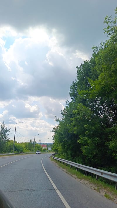 The image shows a view from a vehicle traveling on a paved road. The road curves to the right, with a metal guardrail on the right side. On the left side of the road, there are trees and streetlights. In the distance, there are more trees, some buildings, and a sign. A white car is visible driving on the road in the distance.
The sky is filled with clouds, with patches of blue sky visible. The sun appears to be shining through the clouds, creating bright areas. This suggests it is daytime, likely late morning or afternoon. The lush green trees indicate that it is likely summer.
The overall scene is one of a suburban or rural landscape, with a road leading through a wooded area. The mood is calm and serene. There is no visible text in the image.