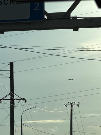 An outdoor medium shot captures a hazy sky with a silhouette of an airplane in flight. In the foreground, a dark, angled structure partially obscures the view of overhead wires. Below this, a dark vertical pole with crossbars and insulators supports multiple wires, with another pole visible to the right. A streetlamp extends from the left side of the frame. A blue sign with the number "2" is visible at the top of the image, with partial Cyrillic text. The scene appears to be in Novomar'evskaya, Russia, given the context provided.