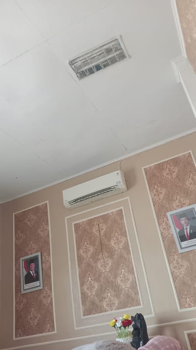 The image shows an interior scene, likely a room in Pulorayeuk, Indonesia. Dominating the upper portion of the frame is a white ceiling with a recessed fluorescent light fixture. Below the ceiling, on the wall, is a white air conditioning unit. The walls are decorated with a patterned wallpaper in shades of beige and brown, framed by white molding. Two framed portraits of men in formal attire are visible on the walls. In the foreground, a vase with a colorful arrangement of flowers is placed on a surface, next to a dark backpack and what appears to be a person's head wearing a pink head covering. The overall impression is of a decorated indoor space.