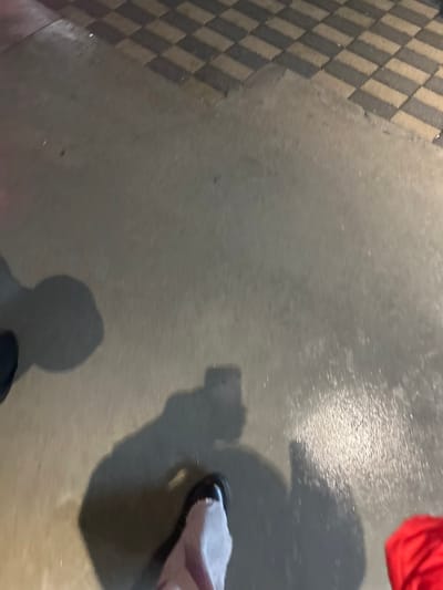 A high-angle shot reveals a checkered tile pattern on the left, transitioning to a smooth, gray surface. A shadow of a person holding a phone is cast on the gray area. Part of a red garment appears in the upper right corner. A foot wearing a pale shoe is also visible, further implying a person standing on the surface.