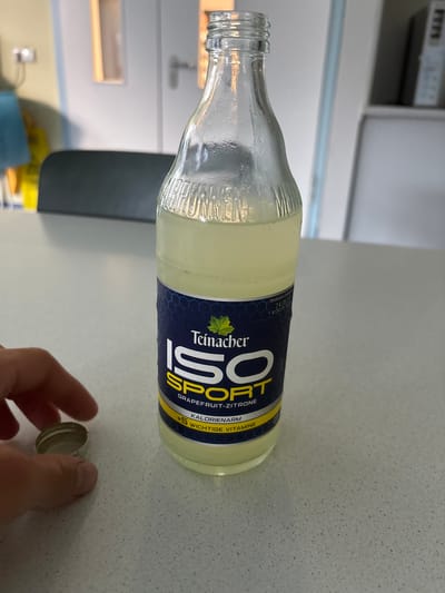 The image shows a glass bottle of Teinacher ISO SPORT Grapefruit-Zitrone drink on a speckled countertop. The bottle is partially full of a light yellow liquid and has a blue and yellow label. Text on the label includes "Teinacher ISO SPORT", "Grapefruit-Zitrone", "KALORIENARM", and "+5 WICHTIGE VITAMINE". A silver bottle cap is held by a person's hand in the foreground, to the left of the bottle. The background is blurred but appears to be an indoor setting with furniture and what looks like a door. There is no visible indication of the city of Mannheim, Germany, in the image.
