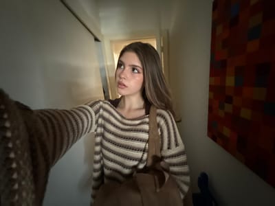 The image displays a young female subject positioned in what appears to be an indoor hallway or entrance area. She is captured from a slightly low angle, indicating a self-portrait. The subject, with medium-length light brown hair and light complexion, is looking upwards and towards the right of the frame, her expression neutral. She is dressed in a horizontally striped knitted sweater in shades of brown and off-white, worn over a dark brown undergarment. A light brown, possibly suede, tote bag is carried over her left shoulder, resting against her front. Her right arm, covered by the sleeve of the same knitted garment, extends into the foreground towards the left.

The immediate environment consists of a plain light-colored wall on the left, featuring a horizontal track or rail near its top edge. In the background, an open doorway reveals a brighter, unidentifiable space beyond. The wall to the right of the subject is adorned with a large, rectangular piece of abstract art, characterized by a grid pattern of multicolored squares, predominantly in shades of red, orange, and yellow. A dark, indistinct shadow or object is present on the floor directly beneath the artwork. Lighting within the scene is uneven, creating contrasts between illuminated and shadowed areas. There are no discernible architectural features, landmarks, or specific visual identifiers within the image to confirm the location as Porto Alegre, Brazil.