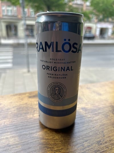 A can of Ramlösa Original mineral water sits on a wooden table. The can is silver and blue with the Ramlösa logo and text in black. The can is in focus, and the background is blurred. The location is a restaurant or cafe in Malmö, Sweden (55.597, 13.009), based on the image's geolocation data. The text on the can reads "Kolsýrat Naturligt Mineralvatten Original Från Ramlösa Hälsobrunn" and features a seal with a man's profile image. The can is sitting on a wooden table, and the rest of the environment is blurred.