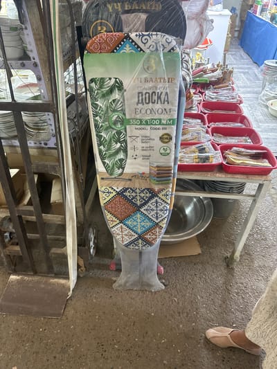 This is a medium shot, eye-level, horizontal, outdoor photograph of a market stall in Kara-Suu, Kyrgyzstan. In the foreground, an ironing board with a patterned cover is leaning against a metal cart. The ironing board has a plastic wrap around it. To the left of the ironing board is a metal shelving unit stacked with white bowls and plates. In the background, a table is laden with red plastic baskets filled with cutlery. Behind the table, there are various items including metal buckets and what appears to be a white chest of drawers. The ground is concrete with scattered debris. A person's feet are visible in the lower right corner of the frame, wearing a light-colored flat shoe. Another person's feet, wearing a pink shoe, are visible near the bottom center of the ironing board. The overall scene depicts a busy market environment.