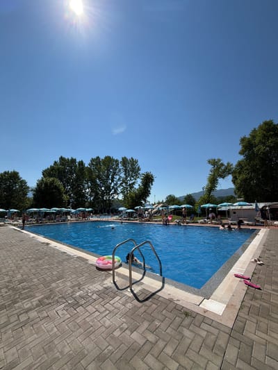 This image depicts a bright, sunny day at an outdoor swimming pool complex, likely located in Simitli, Bulgaria. The foreground is dominated by a paved area made of grey interlocking bricks, which leads to the edge of a large, rectangular swimming pool filled with clear blue water. A set of stainless steel pool stairs with handrails is partially submerged in the water on the right side of the frame. Near the stairs, a colorful inflatable ring floats on the surface of the water.
On the opposite side of the pool, numerous lounge chairs with open umbrellas are arranged, indicating an area for sunbathing and relaxation. Several people are visible around the pool, some are in the water swimming or wading, while others are seated or standing on the deck. The pool area is bordered by lush green trees and foliage, with mountains visible in the background under a clear blue sky. The sun is high in the sky, casting bright sunlight and creating reflections on the water's surface. Scattered on the paved area near the pool's edge are a few pairs of flip-flops. The scene suggests a recreational environment, possibly a public pool or a resort.