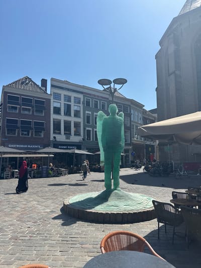 In the city of Zwolle, Netherlands, a public square is depicted on a sunny day. The central focus is a tall, translucent green sculpture of a human figure with wings, resembling an angel, standing on a circular, layered base. In the background, several multi-story buildings line the square. One building, to the left, has dark brickwork and awnings over its windows, with a sign for "GASTROBAR MAXIM'S". Adjacent to it, a white building displays the text "Koning David" and below that, "DOUWE EGBERTS". Further to the right, a large brick church with arched windows dominates the skyline. The ground is paved with cobblestones. Several outdoor seating areas with tables and chairs, some covered by umbrellas, suggest cafes or restaurants. A few people are visible: a person in a red headscarf and black attire walks away from the camera, another person walks towards the right, and others are seated at tables. A row of parked bicycles is present on the right side of the square, and two streetlights with circular tops are positioned near the sculpture.