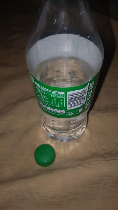 A partially filled clear plastic bottle with a green label is centered in the frame, sitting on a textured surface. A green plastic cap rests beside the bottle on the surface. The bottle's label has nutritional information and the words "RECYCLE ME" printed on it. The liquid inside the bottle appears clear. The background is dark and indistinct. There is no indication this picture was taken in Enugwu-Ukwu, Nigeria.