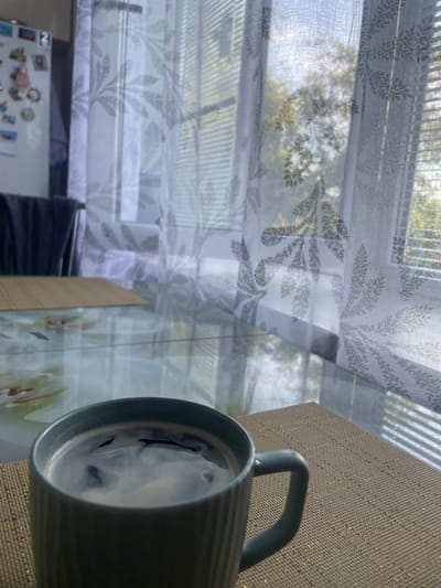 A mug of coffee sits on a table in front of a window with sheer curtains. The curtains are white with a repeating pattern of gray leaves. Outside the window, trees are visible, and sunlight streams through, creating a soft, diffused light. The table appears to be made of glass and has a placemat under the coffee mug. In the background, a refrigerator is visible, adorned with various magnets and pictures. A dark chair is also in the background, partially obscured by the curtains. The scene suggests a peaceful moment at home. The location is not specified to be Ardón, Russia.