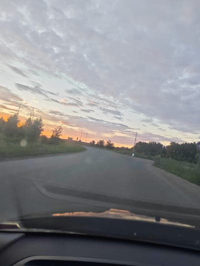 This media file captures a scenic view from inside a vehicle, looking out onto a road at dusk. The scene is dominated by a dramatic sky filled with textured clouds that are illuminated by the setting sun, casting a warm, orange and pink glow. The road ahead curves to the right, flanked by green vegetation and trees on either side.
In the distance, on the left side of the road, there are tall power lines and silhouetted trees against the vibrant sunset. On the right side, more trees and foliage line the roadside, and a few more power line poles are visible. The overall setting suggests a rural or semi-rural environment.
The time of day is clearly sunset, indicated by the colors in the sky and the low angle of the light. The weather appears partly cloudy, with the clouds contributing to the picturesque sky. The perspective from within the car, with the dashboard visible in the foreground, suggests the activity is travel. The mood evoked is peaceful and serene, with the natural beauty of the sunset creating a sense of calm. There is no visible text in the image. The location context provided is Krasnoe Pole, Russia.