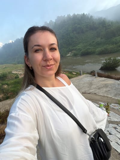 A woman with long brown hair, wearing a white top and a black crossbody bag, is taking a selfie outdoors. She is smiling and looking at the camera. Behind her, a hazy landscape of green hills and a river is visible. The scene appears to be in the city of Nam Khanh Vinh, Vietnam, suggested by the dense vegetation and the style of the surrounding terrain. The ground in the foreground is made of stone and concrete. A plastic bottle of water is also visible to the right of the woman.