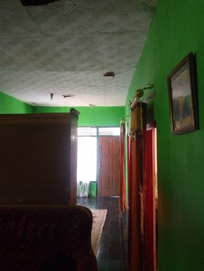 The image depicts the interior of a room, likely a hallway or living area, in a residential building located in Sukamanah, Indonesia. The walls are painted a vibrant green. The ceiling is made of patterned tiles and shows signs of wear, with some visible cracks and damage. A single light bulb hangs from the ceiling, illuminating the space.

On the left side of the frame, a large, dark brown wooden cabinet or wardrobe dominates the scene. In the foreground, the top of a dark red, upholstered sofa is visible. A patterned rug is laid out on the dark tiled floor, extending towards a doorway.

The doorway, on the right side of the hallway, features a wooden door with multiple panels and is open, allowing light to stream in. Beyond the door, a window with horizontal blinds is visible, and sheer white curtains hang. Two sets of curtains, one in a reddish-brown hue and another in a lighter brown, are draped from rods on either side of the doorway, partially obscuring the view further inside.

On the green wall to the right, a framed picture hangs. The frame appears to be made of dark wood, and the picture within depicts a landscape or still life. A decorative metallic lamp with a golden finish is mounted on the wall above the curtains. The overall impression is of a simple, lived-in home.