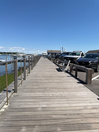 The image captures a wooden boardwalk extending into a harbor on a sunny day in the Village of Sag Harbor, United States of America. The boardwalk, made of weathered gray planks, is bordered on the left by a metal railing and lush green grass, and on the right by a parking area. Several boats, including white yachts, are docked in the calm, blue water to the left of the boardwalk. In the background, a few buildings, including a white structure resembling a small house and another with a dark roof, are visible. Parked cars, including a dark SUV, a black pickup truck, and a white truck with emergency lights, are situated along the right side of the boardwalk. A single, empty wooden bench is placed on the boardwalk, facing the water. The sky above is a clear, bright blue, with no clouds visible.