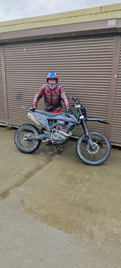A man wearing a blue, white, and red helmet and a red jacket is posing next to a black dirt bike on a wet, gray concrete surface. The background is a brown corrugated metal wall with the number "1/12" visible in the top right corner. It is raining. The tires of the dirt bike are muddy. Based on the presence of water and dirt, the environment is likely outdoors. The location is Kazachiy Brod, Russia.