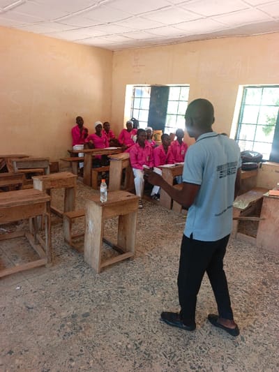 The image displays a classroom interior located in Mada, Nigeria. A male individual stands in the foreground, facing away from the viewer, addressing a group of students. He wears a light blue short-sleeved polo shirt with a dark logo on the back, black trousers, and dark slip-on shoes; his right hand is slightly raised, holding a small white item. Approximately twelve young individuals, predominantly dressed in matching bright pink long-sleeved shirts, are seated at individual wooden desks and chairs in rows in the midground. Some female students wear white head coverings. Their attention is directed towards the standing individual. The classroom walls are light brown with visible white chalk or paint markings, including phrases such as "Good morning," "Cool P," and "Gray Light." The ceiling features light-colored square acoustic tiles. The floor is composed of speckled terrazzo. Two large, multi-paned windows at the back of the room provide natural light and reveal greenery outside. Several empty wooden desks are present in the left foreground, one of which holds a clear plastic water bottle. A dark bag rests on a desk near the right window.