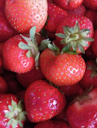 A close-up, overhead shot captures a vibrant pile of fresh, ripe strawberries. The berries are a deep red, glistening with dew or water droplets, and dotted with tiny yellow seeds. Green leafy stems are visible on some of the strawberries, adding a touch of contrast. The focus is sharp on the textures and colors of the fruit, with a shallow depth of field blurring the background. The overall impression is one of natural abundance and freshness. The image appears to have been taken indoors or in a controlled setting, possibly a market or kitchen, given the lighting and lack of environmental context suggesting Hlynytsi, Ukraine.