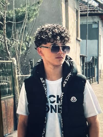 An image depicts a young male standing outdoors in Krivodol, Bulgaria. The subject has dark, tightly coiled curly hair, faded on the sides. He wears large, square-framed aviator-style sunglasses with dark lenses and gold-colored frames. He is dressed in a white short-sleeved t-shirt, which has partially visible dark lettering on the chest, possibly "CK ONE." Over the t-shirt, he wears a black sleeveless puffer vest. The vest features the word "MONCLER" repeatedly printed on the inner collar/hood lining, and a circular Moncler logo patch is visible on the left chest. The subject is looking slightly to his right, away from the camera. The background on the left consists of bare tree branches and a dark metal gate with a wooden panel behind it. On the right, a light-colored building with visible window frames and a roofline is present, indicative of an older architectural style. The scene is illuminated by natural daylight.
