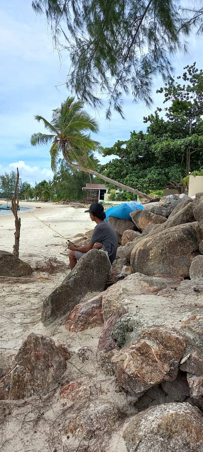 The image shows a man crouching amongst rocks at Chaloklum Beach in Baan Chaloklum, Thailand. The man wears a dark shirt, light-colored shorts, and a dark baseball cap. He is holding an object in his hands and appears to be working on something between the rocks. The beach is visible in the background, with sand and trees. A blue bag is wedged between some rocks behind the man. The sky is partly cloudy.