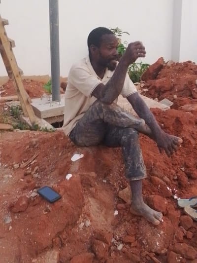 A man with dark skin and a beard sits on a mound of reddish-brown dirt. He wears a light-colored shirt and dirty pants. His hands, feet, and lower arms are also covered in dirt. A cell phone rests on the dirt beside him. A white wall and a pole are visible in the background, along with a ladder and other piles of dirt. The man is gesturing with his right hand as if in conversation.