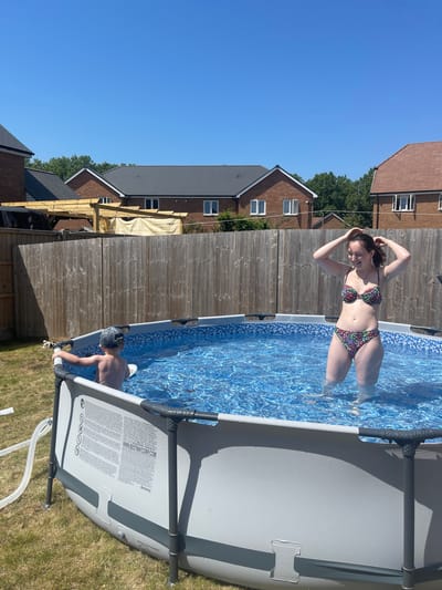 The image depicts a sunny outdoor scene, likely in a backyard, with a blue above-ground swimming pool as the central element. A woman wearing a floral bikini stands waist-deep in the pool, with her hands raised toward her head. A child, possibly a young boy wearing swimming goggles, is also in the pool near the edge, appearing to hold onto the pool's side.
The pool is constructed of gray materials with visible supports. One side panel shows printed information. Hoses are visible near the pool, suggesting its maintenance or filling process.
In the background, there is a wooden fence and residential houses, indicating a suburban setting. The scene occurs in Tonbridge and Malling, United Kingdom.