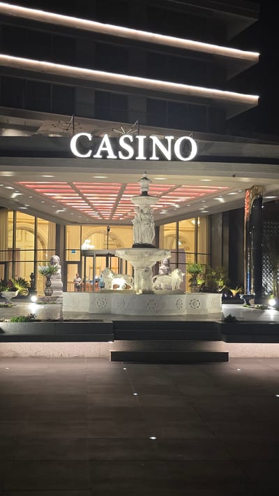 This is a nighttime, eye-level, medium shot of the entrance to a casino in Agios Epiktitos, Northern Cyprus. The casino sign, "CASINO", is illuminated in white block letters against the dark facade of the building. Two horizontal lines of white light run along the upper levels of the building. In front of the casino entrance, a multi-tiered white marble fountain is the central focus. The fountain features sculpted figures, including lions at its base. To the left of the fountain are two stone lion statues. The entrance to the casino has large glass doors and windows, revealing an interior with ornate gold-colored architectural details and a patterned ceiling with red lighting. Several people are visible inside the casino lobby, near the entrance. The ground in front of the casino is paved with large dark tiles, and several small, circular lights are embedded in the pavement. The overall scene is well-lit and presents a grand and somewhat opulent atmosphere.
