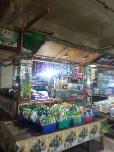 A small sundry store or shop exhibits an array of merchandise. Notable products include numerous bags of "Mama Suka" brand items in blue and green baskets, along with shelves filled with various food products and household items. Advertisements for "Santan" and "Sasa Santan" are prominently displayed, indicating a focus on coconut milk products. The store's sign reads "Toko H. Ujang."