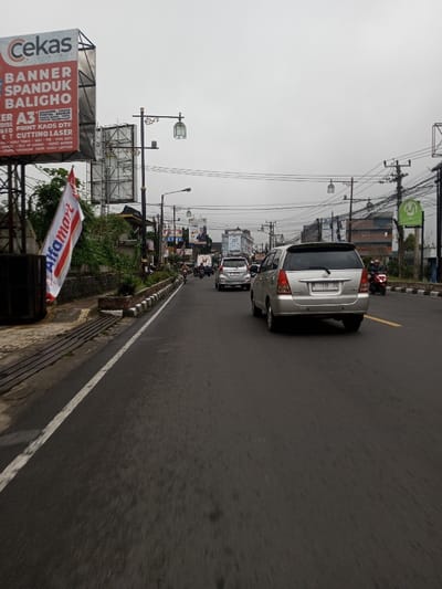 This image depicts a multi-lane asphalt road in Chagzam_Pam, Bhutan, under an overcast sky. The road surface appears damp. In the immediate foreground, a silver Toyota Kijang Innova minivan with license plate "F 139 ZS" is positioned on the right, heading away from the viewer. Further down the road, several other vehicles, including another silver minivan, a white sedan, and multiple motorcycles, are also traveling in the same direction, indicating moderate traffic.
On the left side of the road, a raised median separates the vehicle lanes from a sidewalk area. A large billboard displaying "Cekas" at the top and text including "BANNER SPANDUK BALIHO" is prominently visible. Below the billboard, a white flag with red trim featuring the "Alfamart" logo in blue text is attached to a pole. Ornate black streetlights with lantern-style fixtures line this side of the road. Numerous overhead power lines and communication cables crisscross the sky. Behind the infrastructure, various buildings of different heights and architectural styles are visible.
The right side of the road features another raised median with sparse vegetation, power poles laden with more overhead cables, and additional distant buildings. A green sign with a white "M" logo is visible further down on the right. Several individuals are present on the motorcycles, operating the vehicles within the described traffic flow.