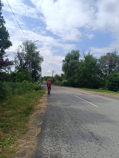 A lone individual wearing a red jacket and a backpack walks along the right side of an asphalt road, facing away from the camera. The road has a white line marking its center and is wet in patches. To the left of the road, there is a strip of green grass and dry, light brown vegetation. Beyond the grass is a dense line of trees and bushes, with some appearing overgrown. In the background, a few utility poles with wires are visible. The sky above is mostly blue with scattered white clouds. The scene appears to be in Ostrogorka, Russia, likely a rural or suburban area given the road and vegetation.