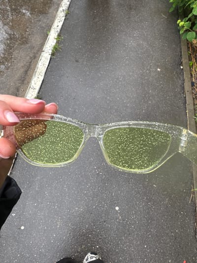 A close-up, slightly downward-angled shot shows a person's hand holding a pair of clear, plastic-framed sunglasses with yellow lenses. The sunglasses are covered in numerous small water droplets, which are also visible on the lenses. The background is a wet, dark asphalt surface, possibly a sidewalk or road, with some grass and vegetation growing along the edge on the right. The lighting suggests an overcast or rainy day. The location is identified as Desnogorsk, Russia.